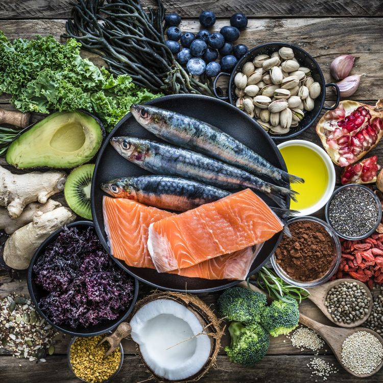 Kompozycja produktów spożywczych na drewnianym blacie: ryby i filet z łososia, awokado, jarmuż, borówki, brokuł, granat, orzechy i nasiona oraz miseczka z oliwą.