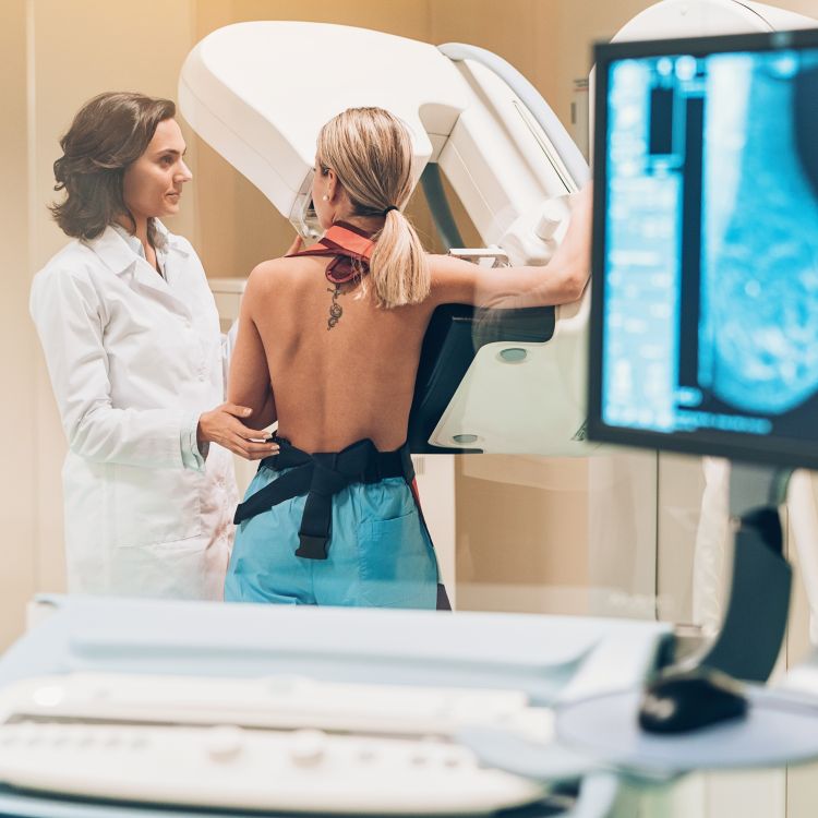 Kobieta w trakcie badania mammograficznego, wspomagana przez lekarkę w białym fartuchu w nowoczesnym gabinecie lekarskim.