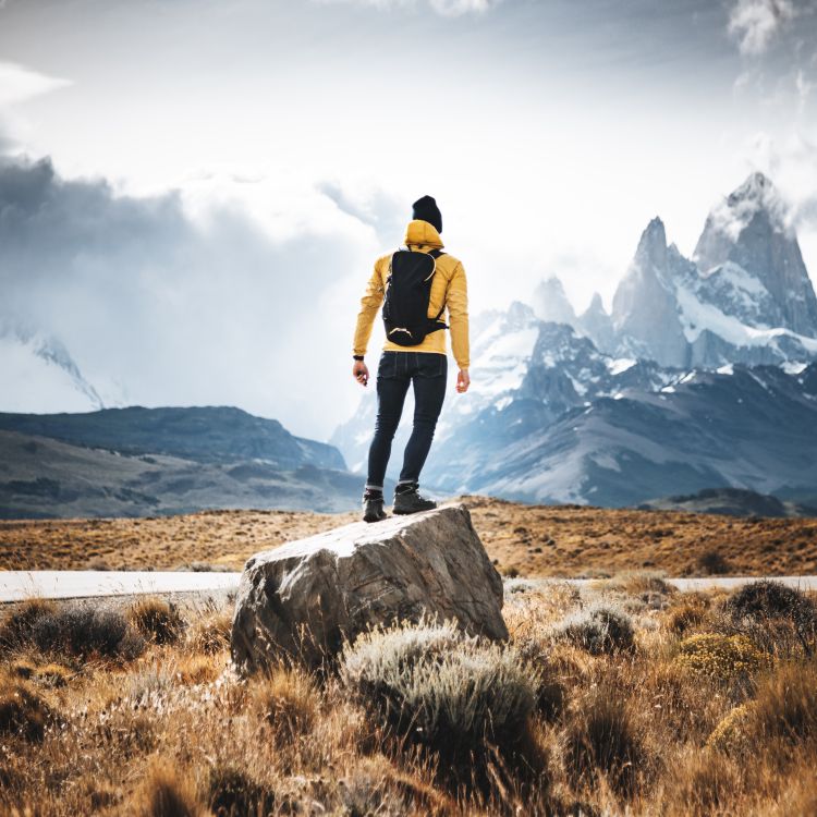 Osoba w żółtej kurtce i z czarnym plecakiem stoi na dużym głazie, patrząc na ośnieżone górskie szczyty w rozległym, surowym krajobrazie.