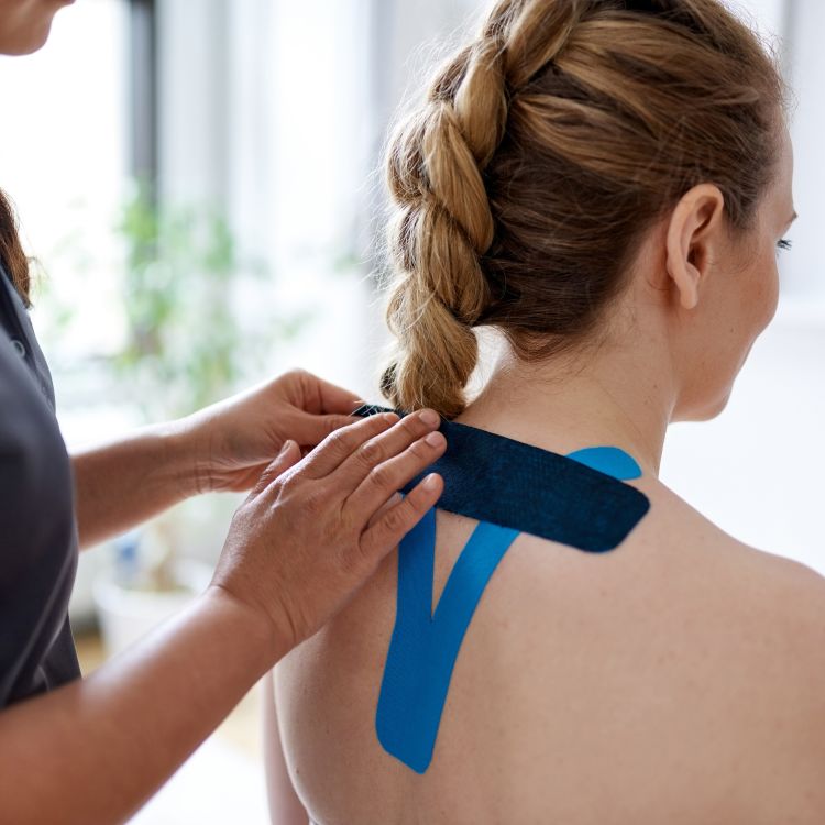 Specjalista przykleja taśmy kinesiotaping na górnej części pleców i barku osoby siedzącej tyłem.