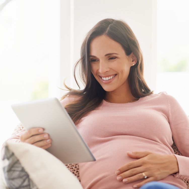 Zdjęcie przedstawia uśmiechniętą, ciężarną kobietę o ciemnych włosach, która z czułością trzyma się za brzuch. Kobieta ma na sobie jasnoróżowy sweter, a na jej palcu widoczna jest obrączka.