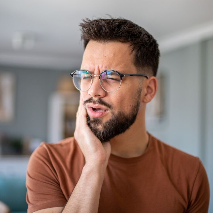 Osoba w okularach dotykająca okolicy żuchwy i policzka w domowym wnętrzu, ilustracja problemów związanych z bruksizmem i napięciem mięśni szczęki.