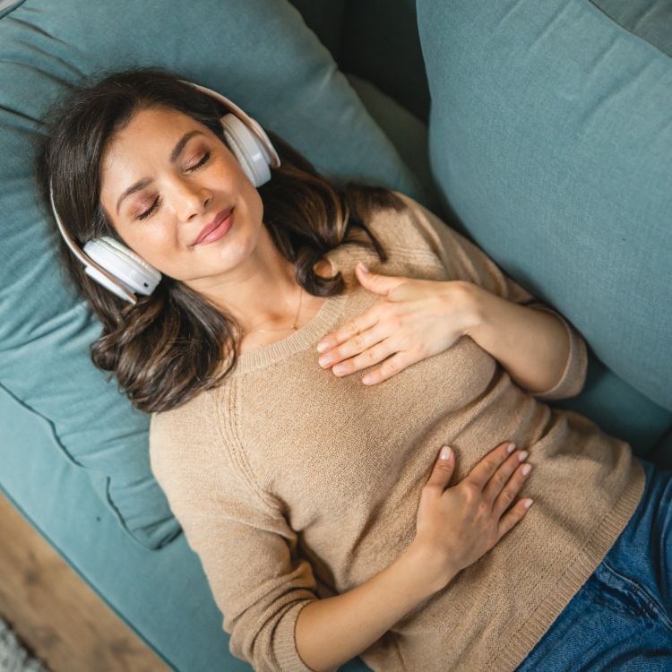 Zdjęcie przedstawia młodą kobietę o ciemnych włosach, leżącą na błękitnej kanapie w stanie głębokiego relaksu. Kobieta ma zamknięte oczy i delikatny uśmiech na twarzy, a na uszach białe, bezprzewodowe słuchawki nauszne. Jej dłonie spoczywają spokojnie na klatce piersiowej i brzuchu, co podkreśla atmosferę wyciszenia i odpoczynku.