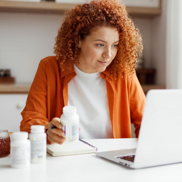 Kobieta w rudych włosach siedząca przy biurku z laptopem trzyma opakowanie suplementu diety; na blacie stoją inne butelki i notes.