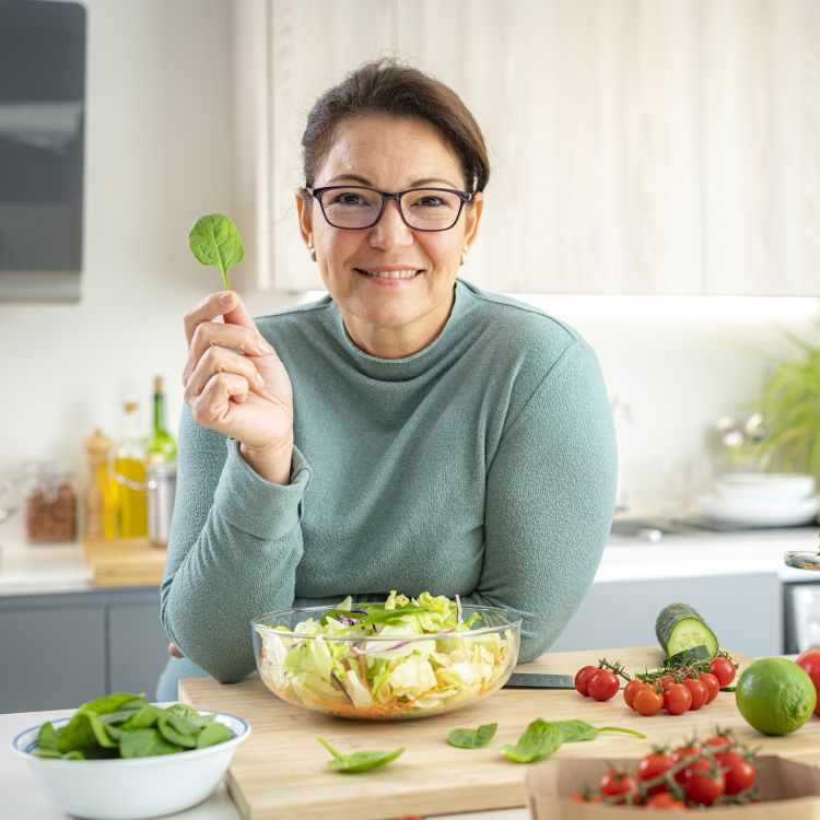 Uśmiechnięta kobieta w okularach i turkusowym golfie siedzi przy kuchennym blacie, trzymając w dłoni pojedynczy liść szpinaku. Przed nią stoi duża miska sałaty oraz świeże warzywa: pomidorki koktajlowe i szpinak.