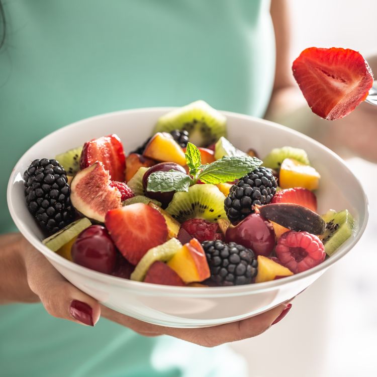 Zbliżenie na dłonie trzymające białą miskę z bogatą sałatką owocową. W składzie widać kawałki truskawek, kiwi, brzoskwiń, figi, a także całe maliny, jeżyny i czereśnie. Na szczycie znajduje się świeża mięta.