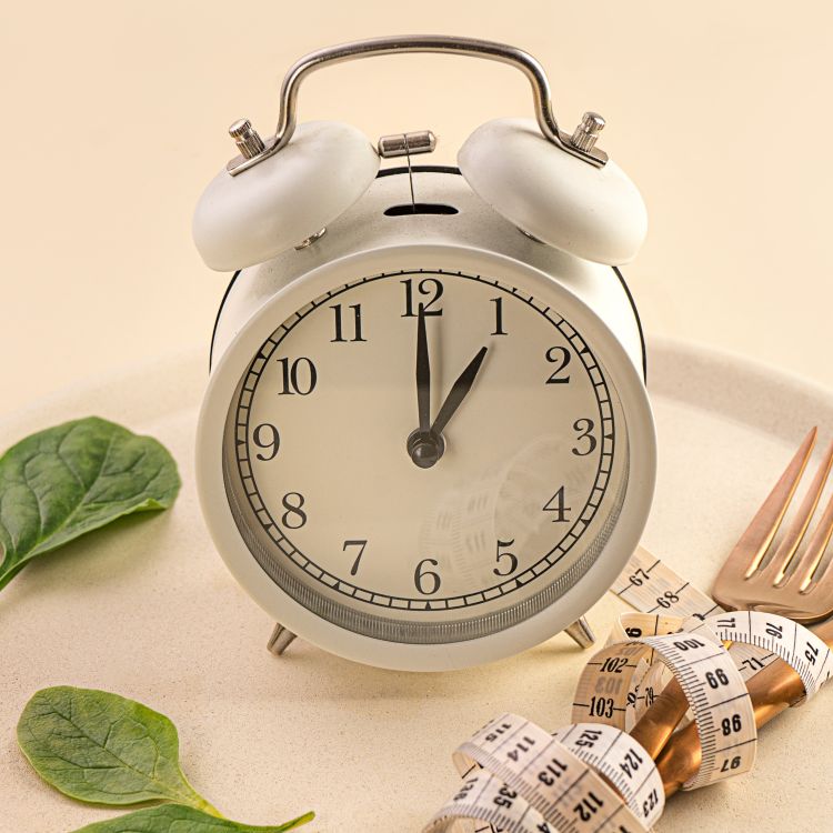 Biały budzik analogowy IKEA DEKAD stojący na białym talerzu, obok zielonych liści szpinaku i zwiniętej miarki krawieckiej. Wskazówki zegara pokazują godzinę pierwszą.