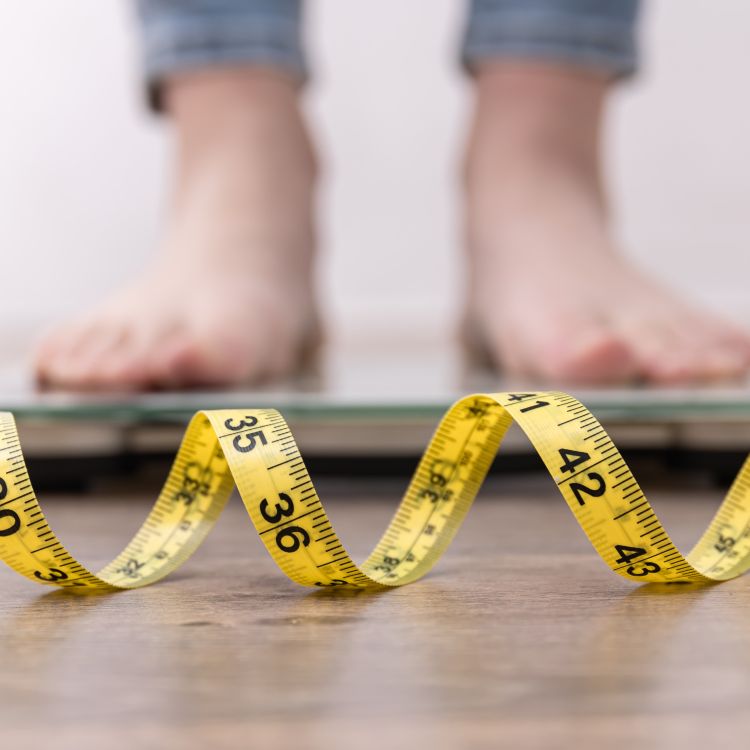 Zbliżenie na stopy stojące na wadze, z żółtą miarką krawiecką ułożoną na podłodze na pierwszym planie.