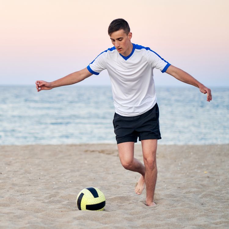 Nastolatek grający w piłkę nożną na plaży.