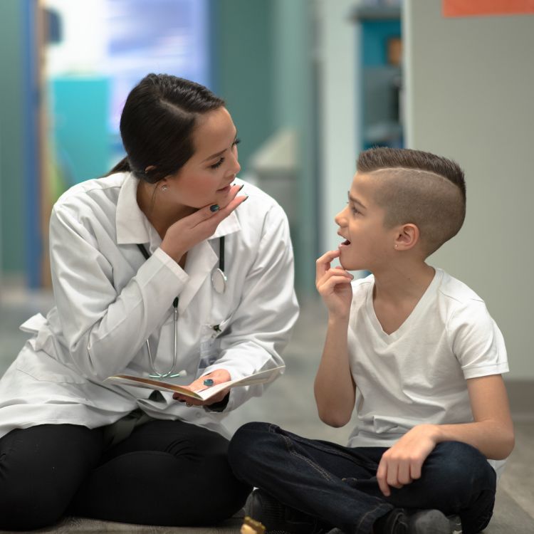 Kobieta w białym fartuchu lekarskim siedzi na podłodze naprzeciwko kilkuletniego chłopca. Oboje patrzą na siebie z uśmiechem i wykonują gesty dłońmi przy swoich twarzach, co sugeruje naukę mowy lub komunikację w języku migowym. Kobieta trzyma na kolanach otwartą książeczkę.