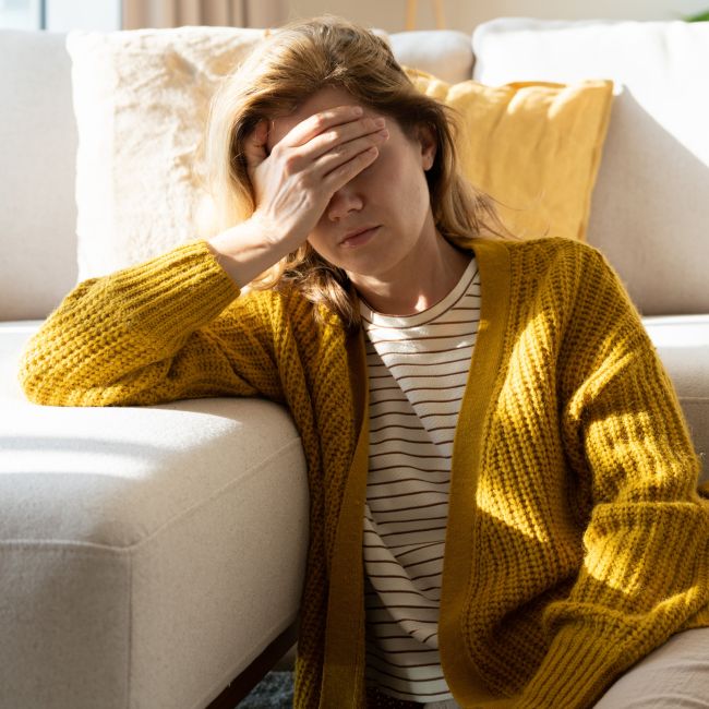 Kobieta w musztardowym kardiganie i pasiastej bluzce siedzi na podłodze, opierając się o białą sofę. Trzyma dłoń na czole i ma zamknięte oczy, co sugeruje silny ból głowy lub migrenę. Pomieszczenie jest jasne, oświetlone ciepłym światłem słonecznym.
