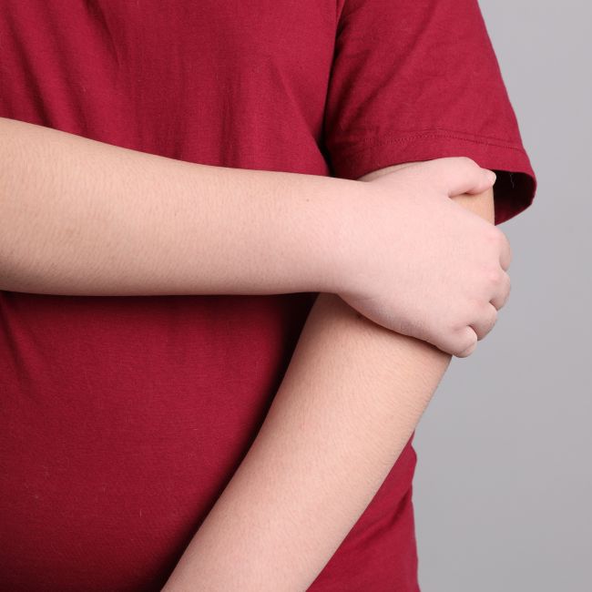 Bliskie ujęcie na tułów i skrzyżowane na brzuchu ręce osoby ubranej w czerwoną koszulkę. Zdjęcie ilustruje tekst dotyczący przyczyn otyłości u dzieci oraz nieprawidłowych nawyków żywieniowych.