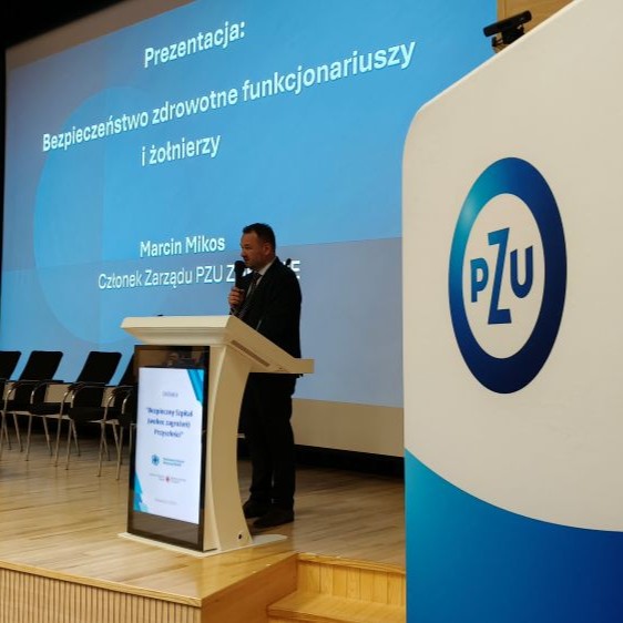 Zdjęcie przedstawia prelegenta, dr. hab. Marcina Mikosa, członka zarządu PZU Zdrowie, przemawiającego za mównicą podczas konferencji. W tle widoczny jest slajd prezentacji o tytule „Bezpieczeństwo zdrowotne funkcjonariuszy i żołnierzy” oraz duże logo PZU Zdrowie na niebiesko-białym tle.