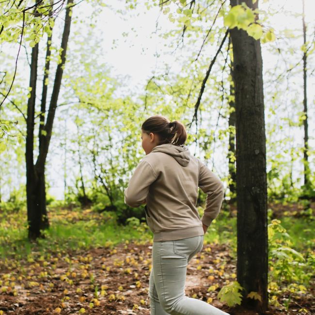 Dziecko z niewielką nadwagą w beżowej bluzie i jasnych spodniach biegnie ścieżką wśród drzew w parku lub lesie.